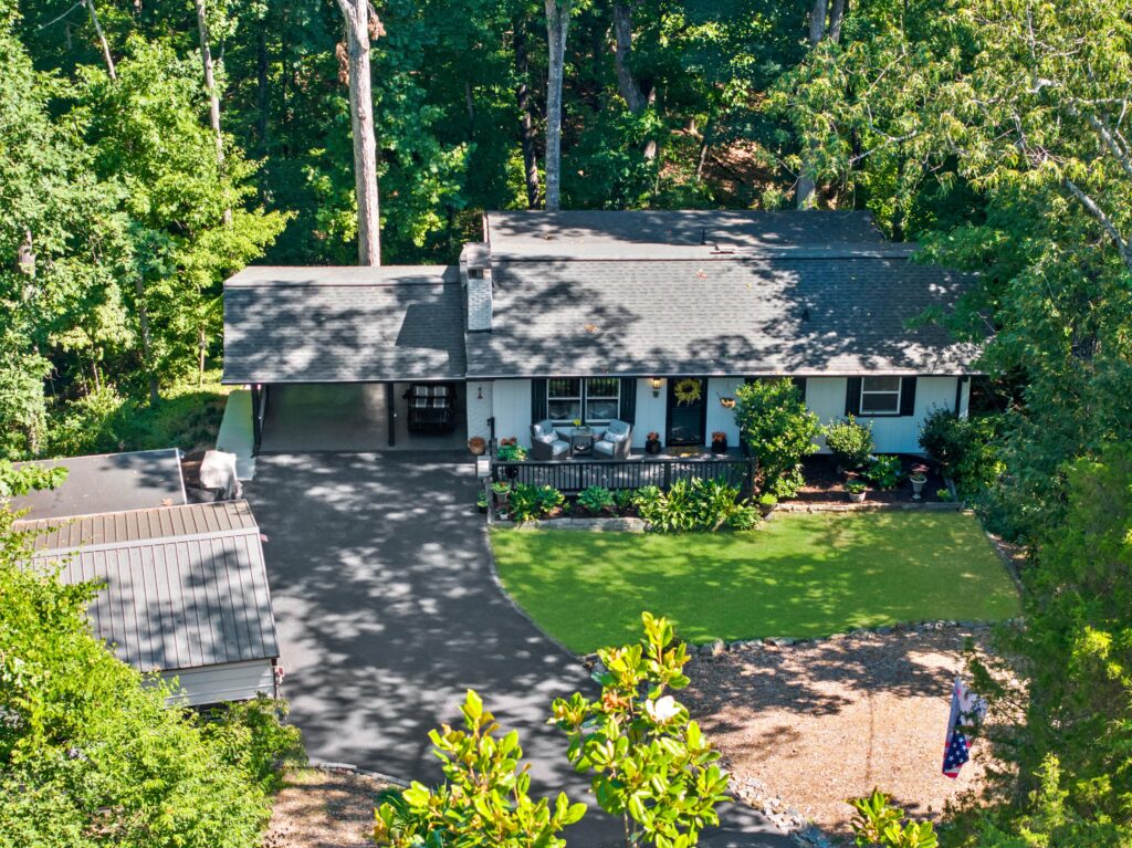 Ranch-style home with carport nestled among tall trees with a mix of lawn and mulch.